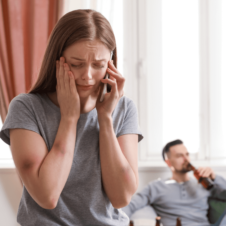 Scared woman on phone with man in background drinking beer.